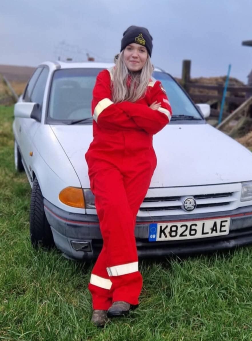 Natalie O'Donnell standing in front of a car in a field wearing a red boiler suit