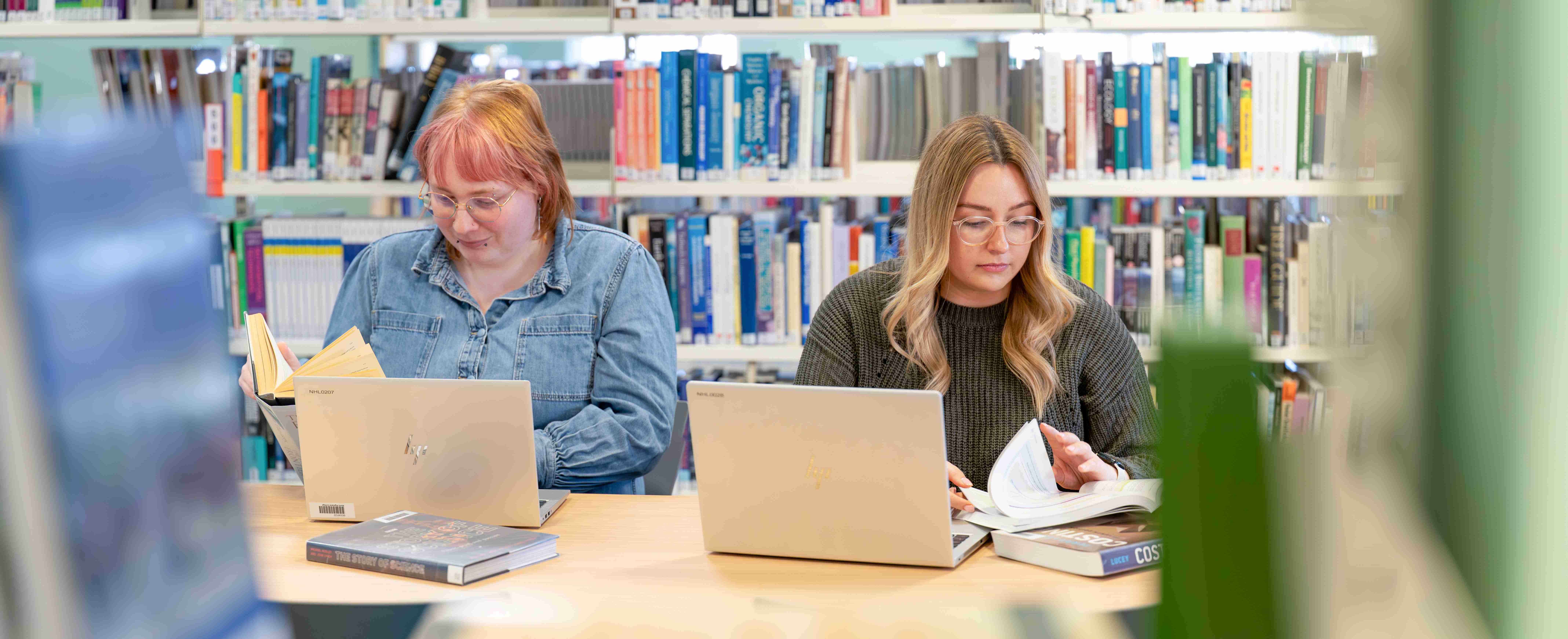 2 students sitting studying in the library with books and laptops and book shelves in the background 2 students sitting studying in the library with books and laptops and book shelves in the background